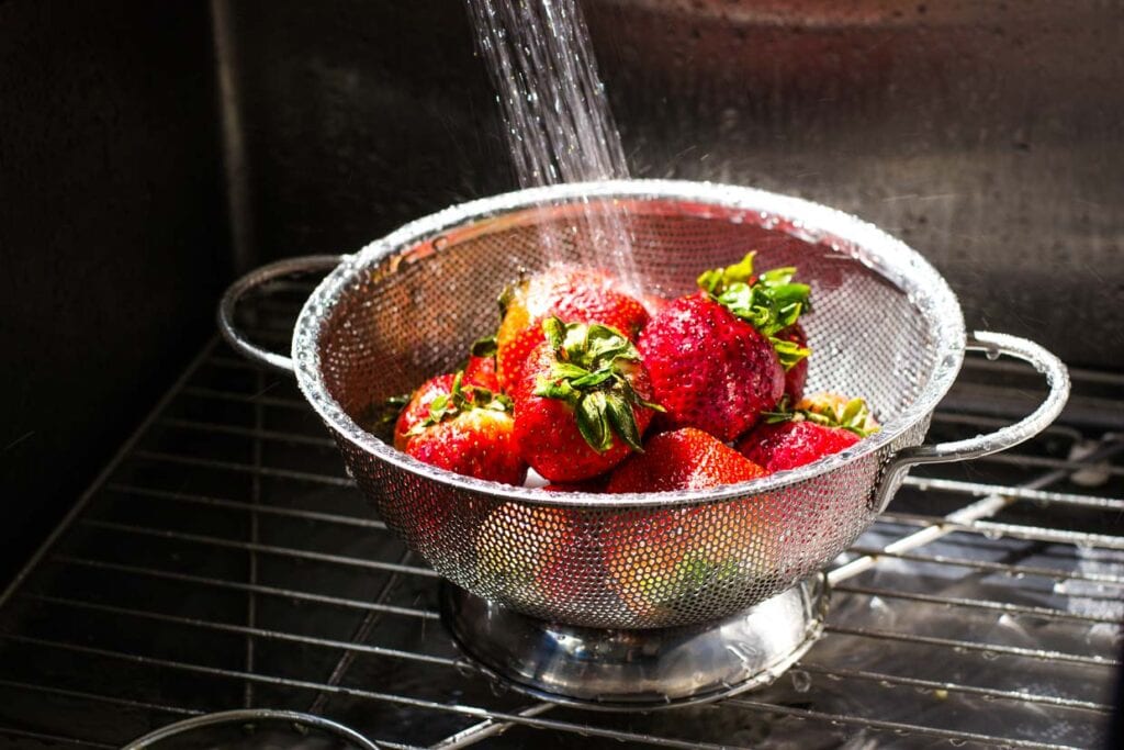 Strawberries, those delightful summer berries, are being rinsed with water in a metal colander placed in a sink.