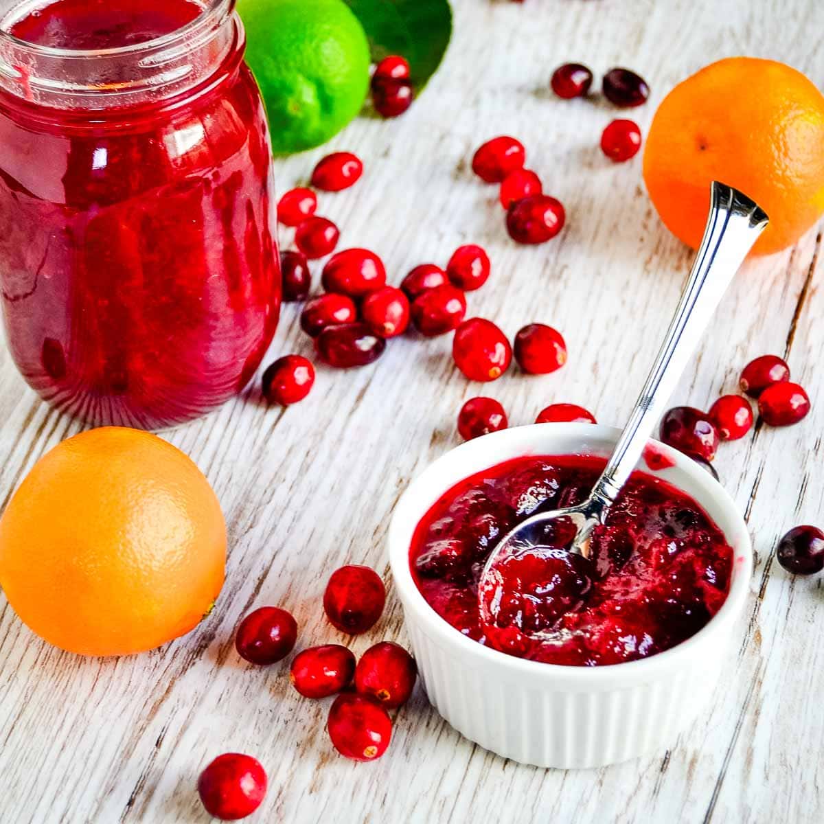 A jar and bowl of cranberry sauce with a spoon, surrounded by fresh cranberries, juicy clementines, oranges, and a lime on a wooden surface.