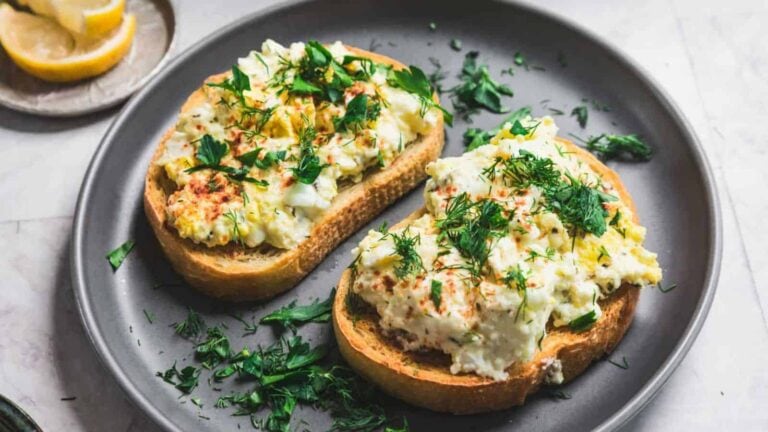 Two slices of bread topped with egg salad and garnished with parsley and dill on a dark plate. Lemon wedges are in the background.