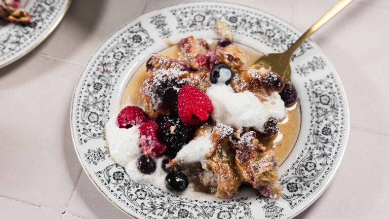 A plate with bread pudding topped with whipped cream, raspberries, blueberries, and powdered sugar. A gold fork is on the plate.
