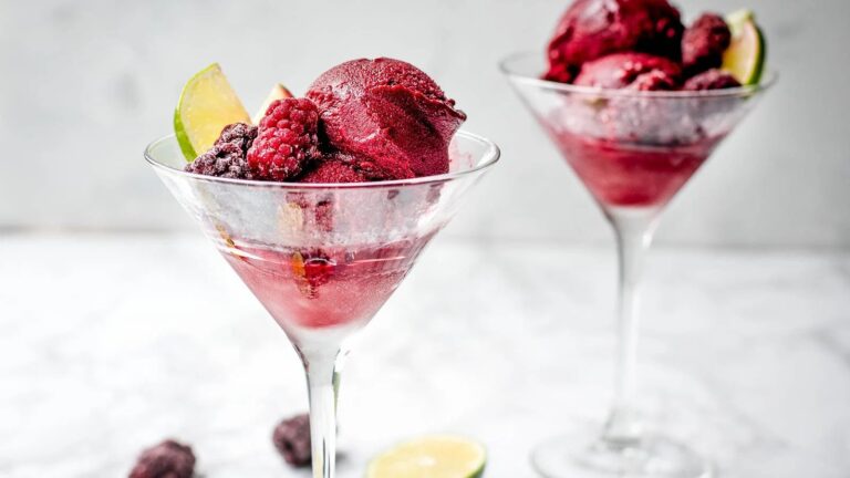 Two martini glasses filled with dark berry sorbet garnished with blackberries, raspberries, and lime slices on a light textured surface.