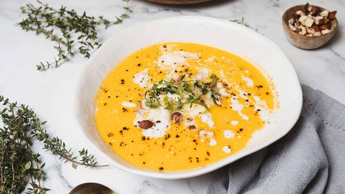 A bowl of creamy orange soup garnished with herbs, chopped nuts, and drizzled cream sits on a marble surface. A small bowl of nuts and sprigs of fresh herbs are nearby, alongside a gray napkin.