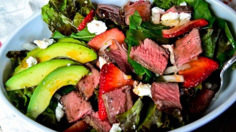 Salad with sliced steak, avocado, strawberries, lettuce, and crumbled cheese in a bowl.