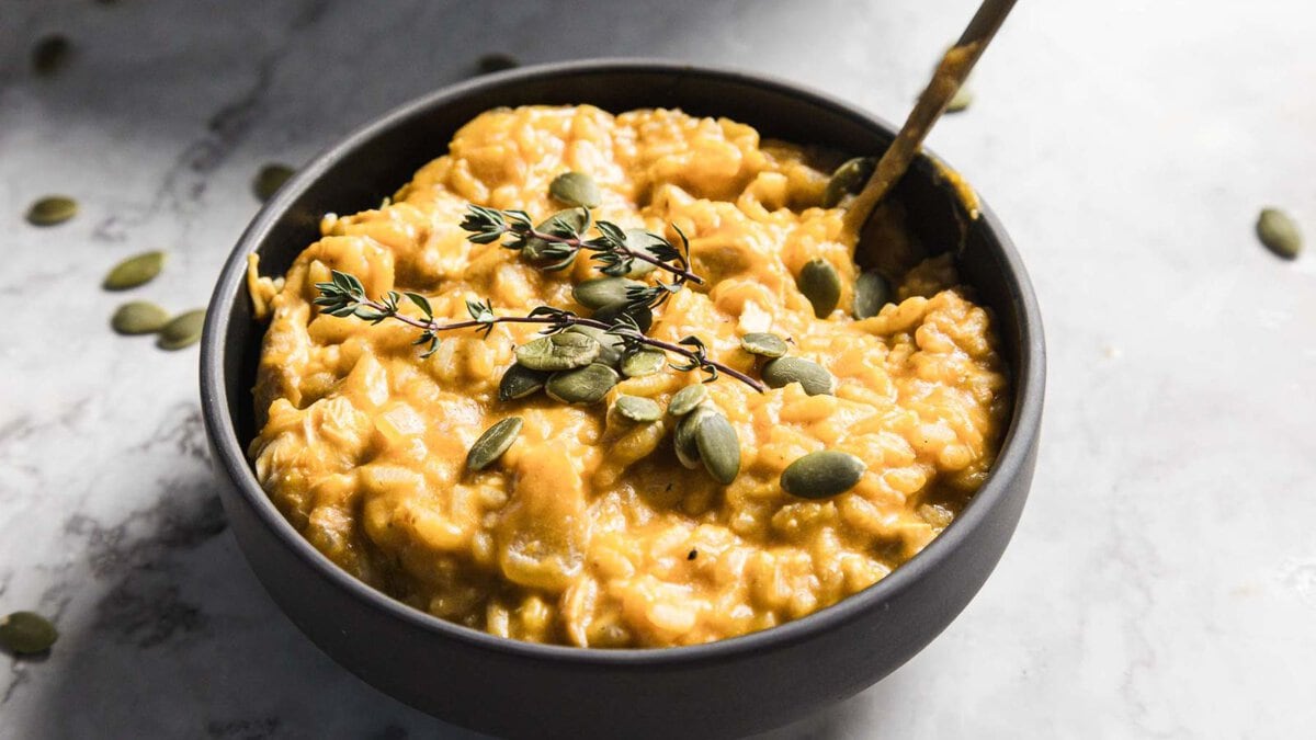 A black bowl filled with creamy, yellow pumpkin risotto. The dish is garnished with fresh thyme sprigs and scattered pumpkin seeds. A spoon rests inside the bowl, and a blurred background shows additional thyme sprigs.