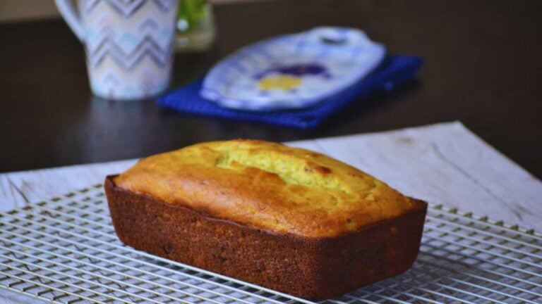 A loaf of golden-brown bread on a cooling rack with a mug and a plate in the background.