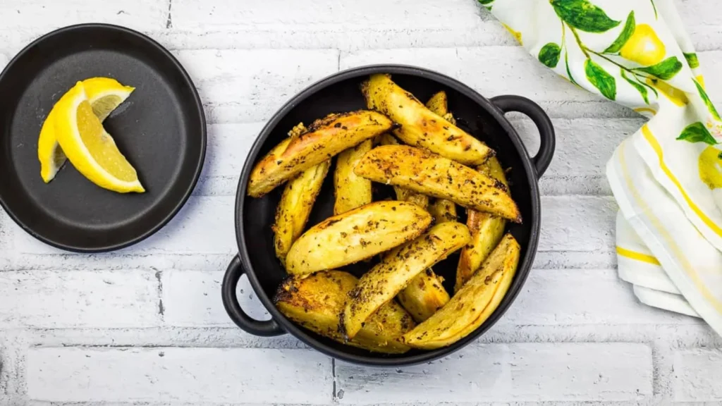A bowl of seasoned potato wedges on a white surface next to a small black plate with lemon wedges. A lemon-patterned cloth is partially visible on the right.