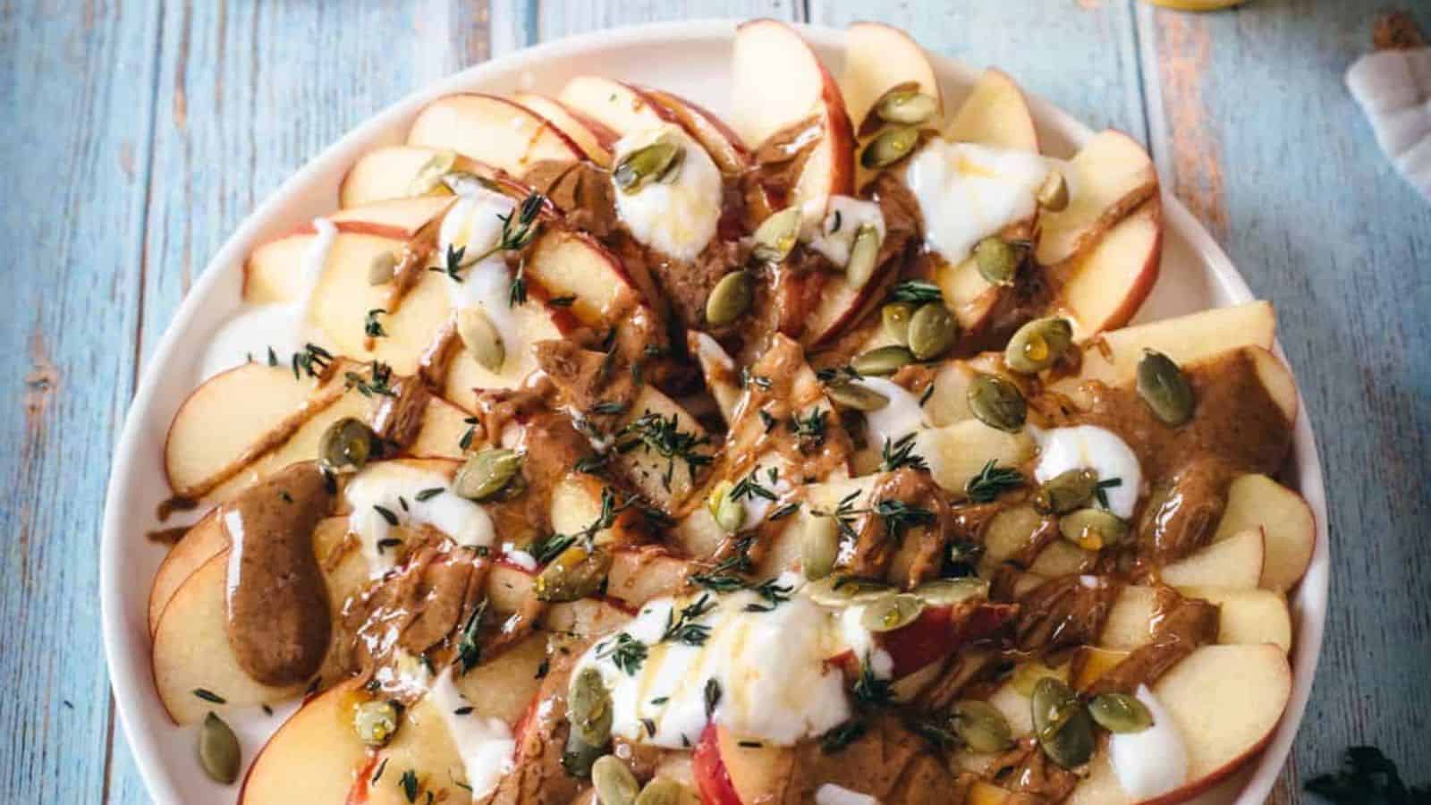 A plate of sliced apples topped with dollops of yogurt, drizzles of almond butter, sprinkled pumpkin seeds, and fresh thyme leaves, displayed on a rustic wooden surface.