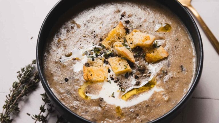 A bowl of mushroom soup topped with croutons, cream, and herbs, with a gold spoon beside it on a white surface.