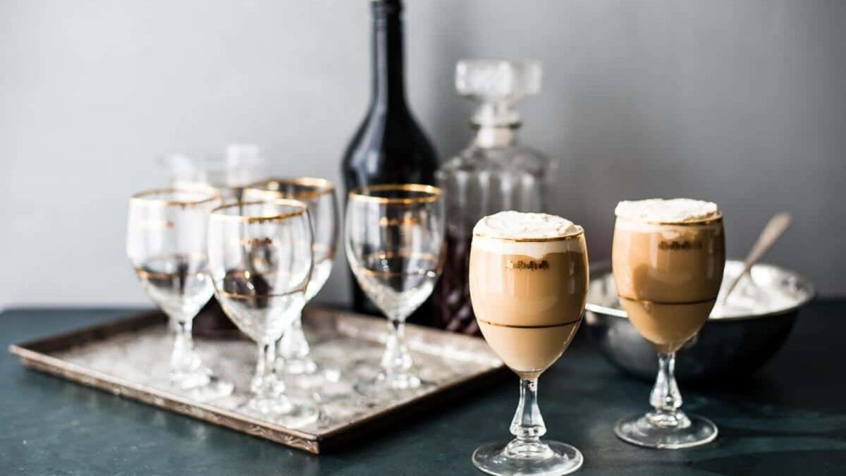 Two glasses of Irish Cream coffee in front of a tray of festive glass and a bar set up for making more.