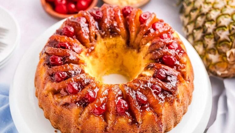 A golden-brown bundt cake topped with glazed cherries and slices of pineapple is on a white plate. Nearby are a bowl of cherries and part of a whole pineapple.