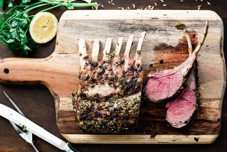 Herb-crusted rack of lamb, sliced, on a wooden cutting board with a lemon half and herbs nearby.