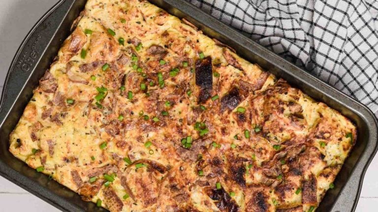 A baked egg casserole with pieces of bread and green onions in a rectangular pan. A checkered cloth is partially visible next to it.