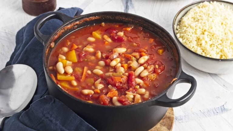 A black pot of bean stew with tomatoes, peppers, and vegetables beside a bowl of couscous on a white surface.