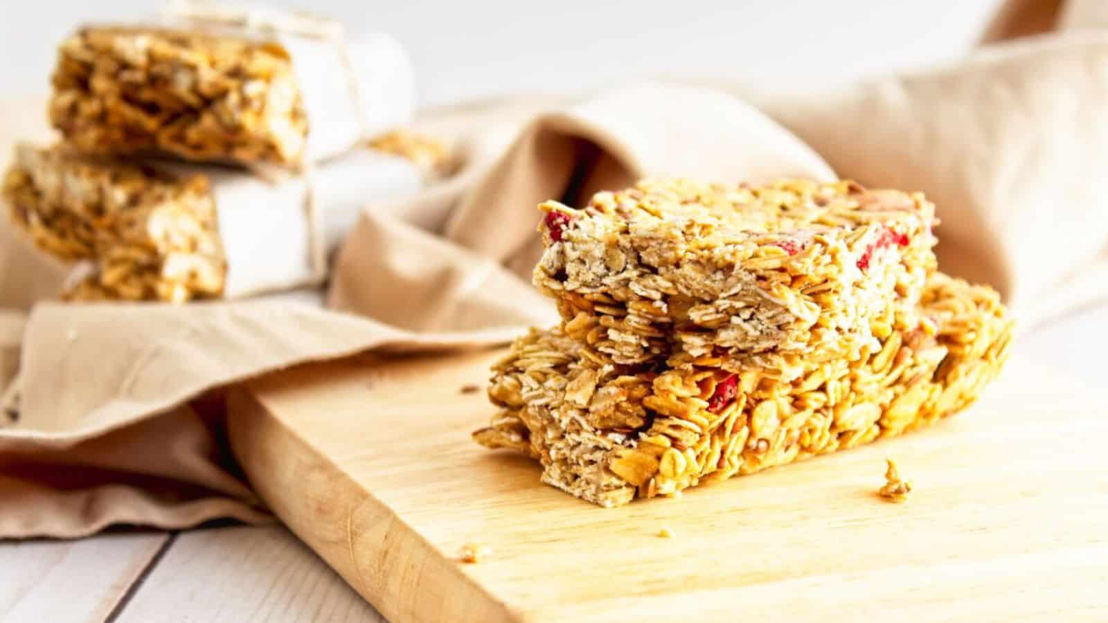 Two granola bars stacked on a wooden board with more bite-sized treats wrapped in parchment paper in the background.