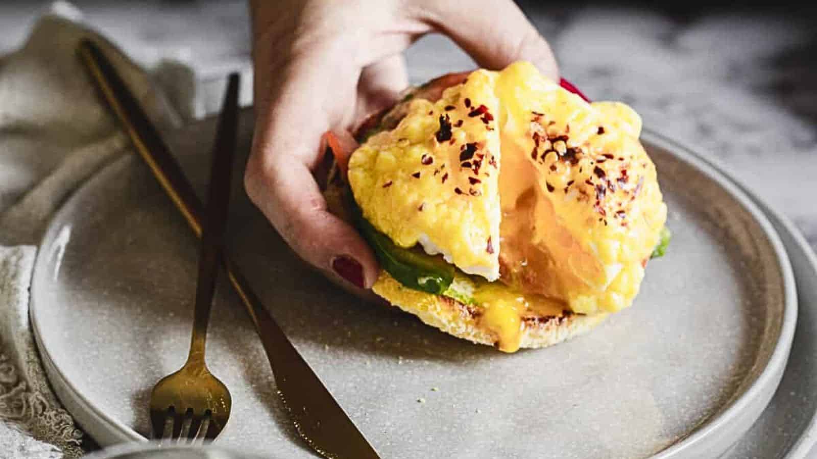 A plate with eggs benedict featuring a poached egg, hollandaise sauce, and avocado slices on an english muffin, garnished with spices, accompanied by gold cutlery. Perfect for Mother's Day recipes.