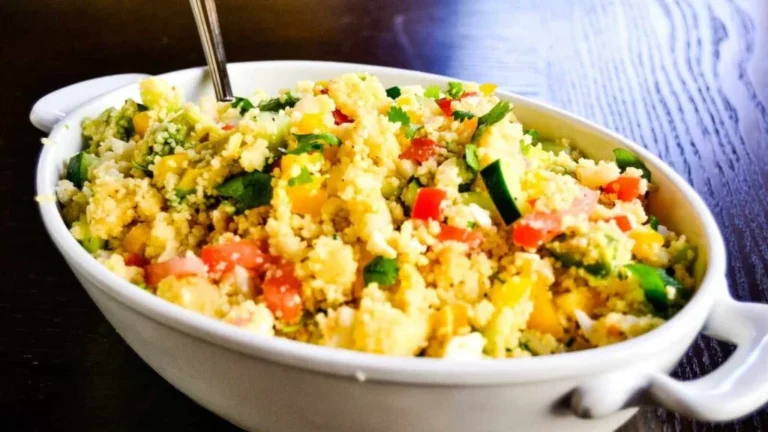 A white bowl filled with couscous salad, containing diced tomatoes, cucumbers, peppers, and garnished with herbs. A spoon is placed in the bowl.
