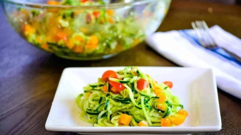 A plate of zucchini noodles with cherry tomatoes and diced yellow bell peppers, served in front of a glass bowl with more salad. A folded napkin and forks are in the background.