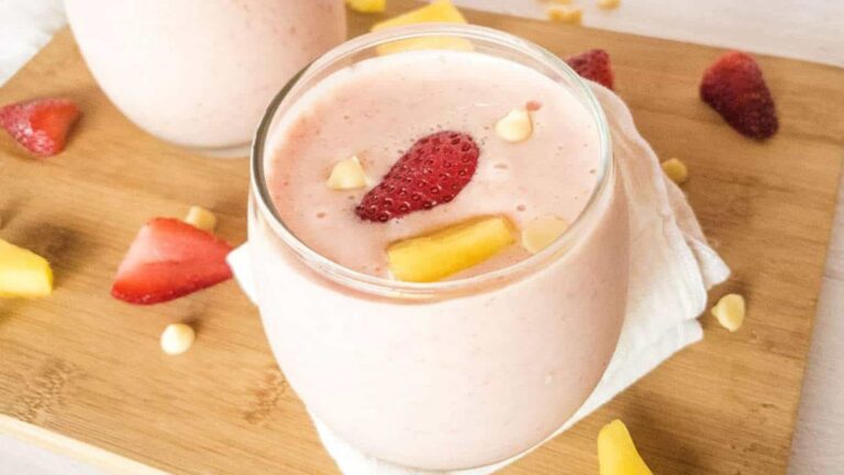 A glass of strawberry mango smoothie on a wooden board, garnished with sliced strawberries and mango chunks, surrounded by scattered fruit pieces.