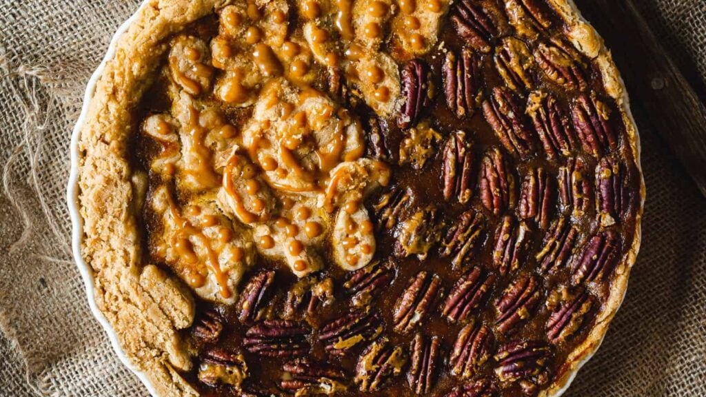 Close-up of a pie with half covered in pecans and the other half topped with caramel and pastry decorations, set on a burlap surface.