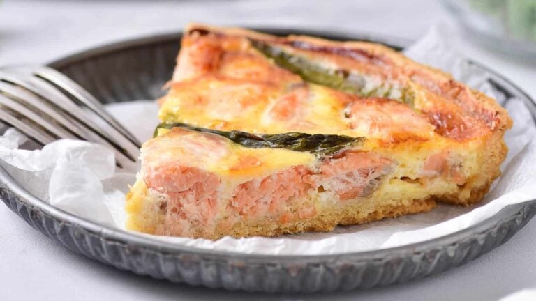 A slice of salmon and asparagus quiche on a paper-lined plate, with two forks beside it, makes for a delightful addition to any AM menu, celebrating breakfasts filled with fresh veggies.
