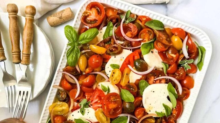 A rectangular dish with sliced tomatoes, mozzarella, red onions, basil leaves, and mixed herbs, next to plates and forks on a white surface.