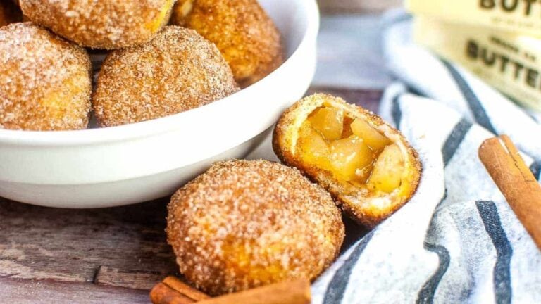 A white bowl filled with sugar-coated dough balls sits beside a halved dough ball revealing an apple filling, with cinnamon sticks and butter in the background—a perfect treat for Mother’s Day and lovers of delicious fruit recipes.