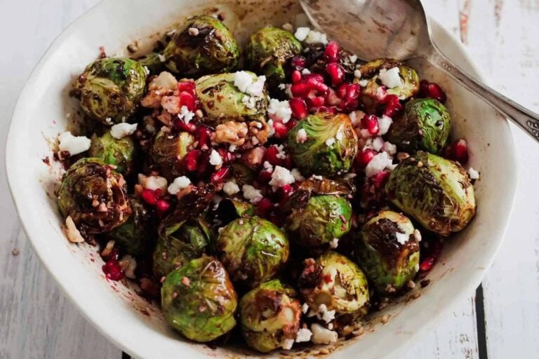 A bowl of roasted Brussels sprouts topped with crumbled cheese, pomegranate seeds, and chopped nuts—one of those classic sides that bring kitchen memories to every meal. Served with a handy spoon for easy sharing.