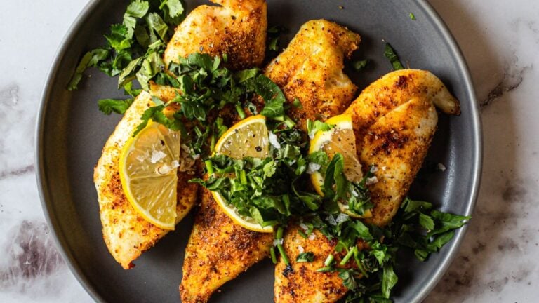 Four seasoned chicken breasts served on a gray plate, garnished with fresh chopped herbs and lemon wedges.