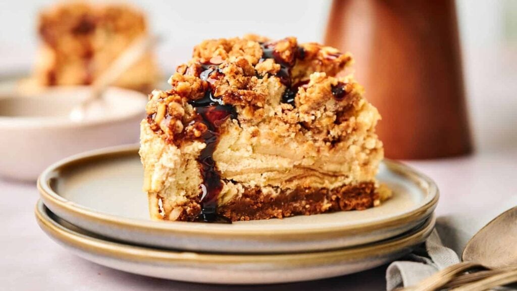 A slice of layered crumb cake with a streusel topping and dark syrup sits on a stacked plate with a fork nearby.
