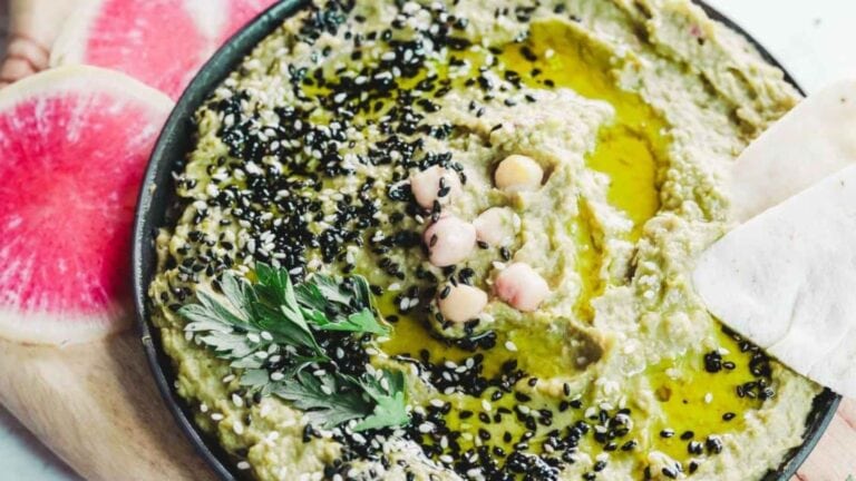 A plate of hummus topped with black sesame seeds, chickpeas, olive oil, and parsley, with sliced watermelon radish on the side.