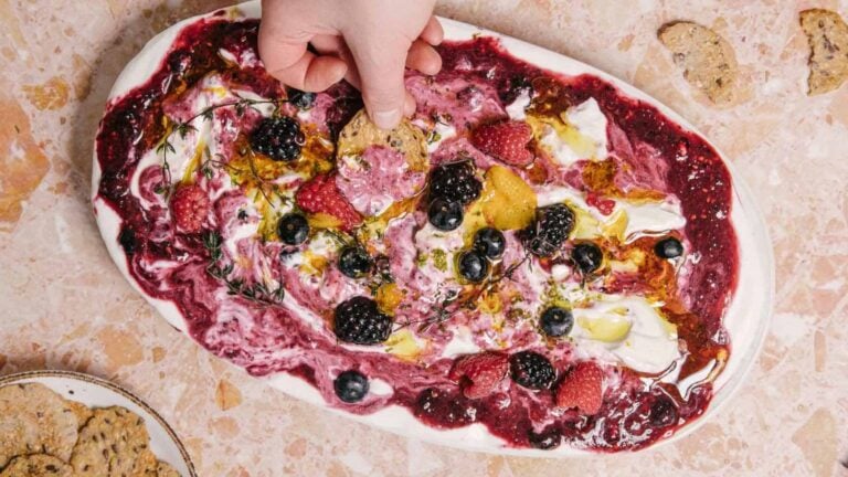 A hand dips a cracker into a platter of whipped cheese topped with a berry sauce, fresh berries, herbs, and olive oil, set on a beige marble surface. A plate with more crackers is visible in the corner.