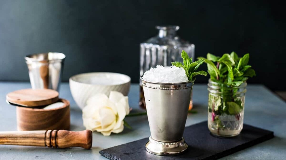 A mint julep in a silver mint julep cup with sprigs of mint and a decanter of bourbon and other ingredients in the background.