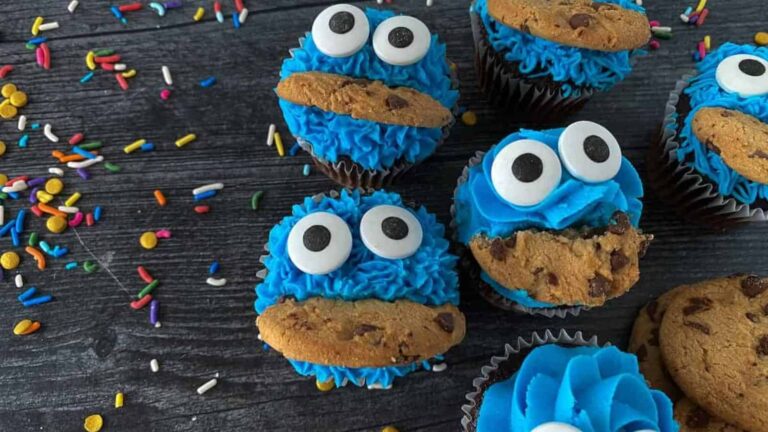 Cupcakes decorated with blue frosting, large candy eyes, and chocolate chip cookies to resemble Cookie Monster, surrounded by colorful sprinkles on a dark surface.
