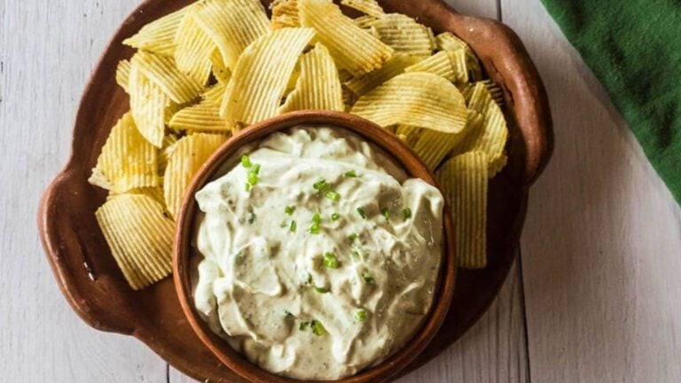 A plate with ridged potato chips and a bowl of creamy dip garnished with chopped green herbs on a wooden surface.