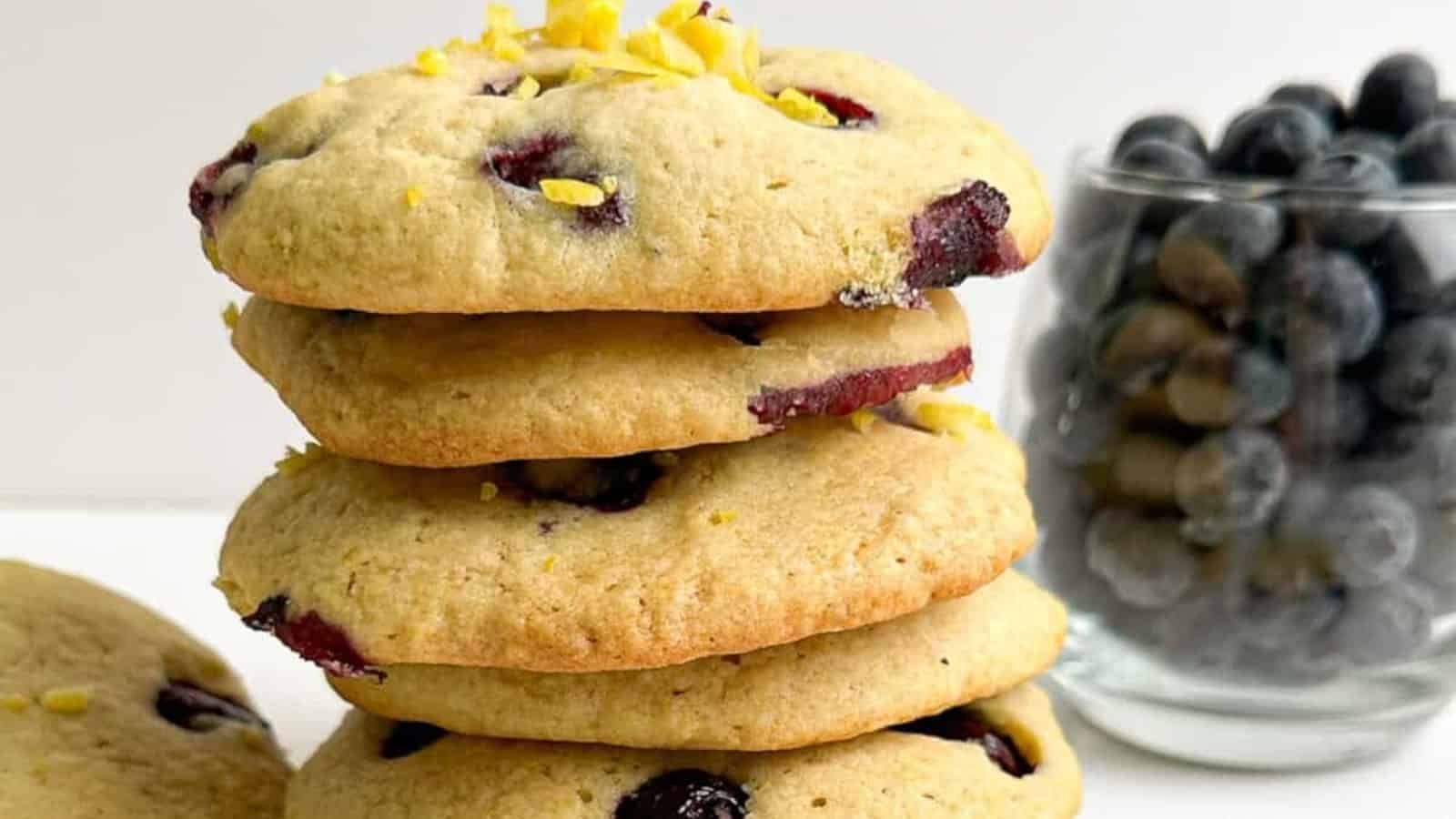 A stack of blueberry cookies with lemon zest on top. A glass jar filled with blueberries is in the background.