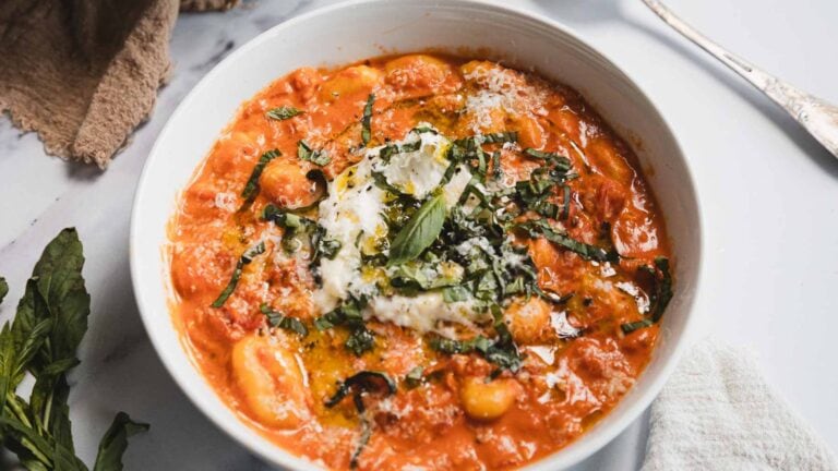 A white bowl filled with tomato-based gnocchi topped with creamy cheese, chopped fresh basil, and a drizzle of olive oil sits on a white surface with a fork, napkin, and basil leaves nearby.
