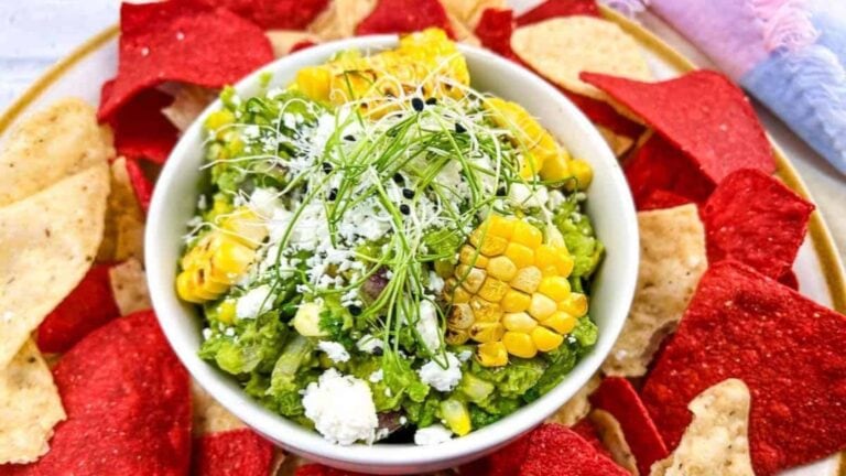 A bowl of guacamole topped with corn, cheese, and sprouts is surrounded by red and white tortilla chips on a plate—an irresistible appetizer perfect for party food platters.