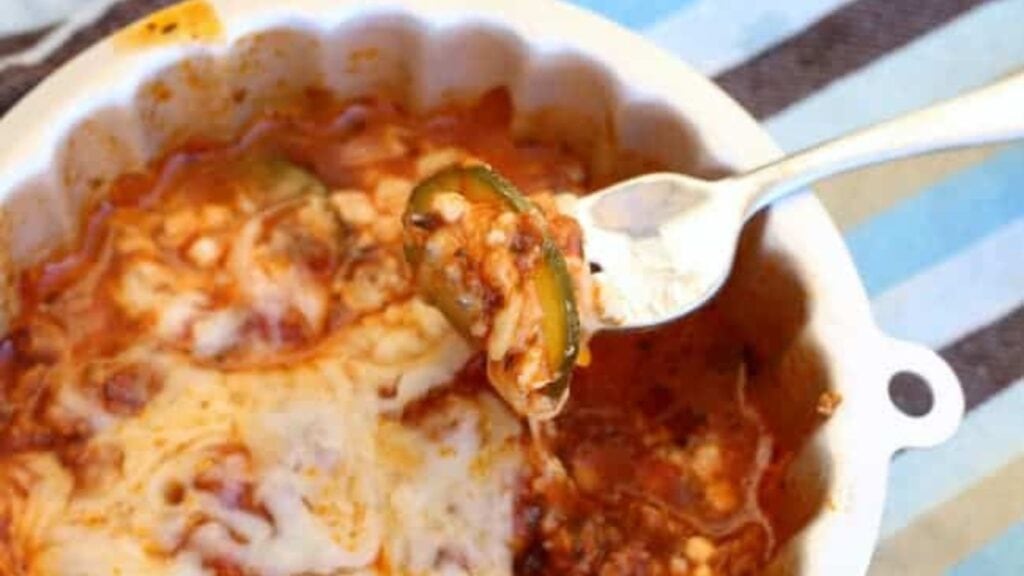 A fork holds a bite of cheesy baked zucchini casserole from a white dish, partially eaten, with sauce and melted cheese visible—a delicious choice for veggie recipes, perfect for your next family potluck.