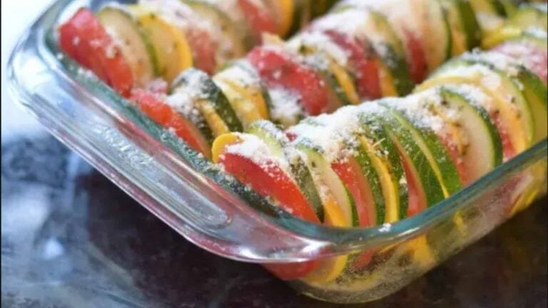 A glass baking dish filled with neatly arranged slices of tomato, zucchini, and yellow squash, topped with grated cheese or seasoning.