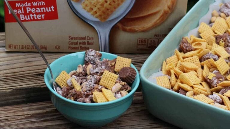 A teal bowl of peanut butter and chocolate cereal mix sits beside a baking dish and a cereal box labeled “Made With Real Peanut Butter” on a wooden surface.