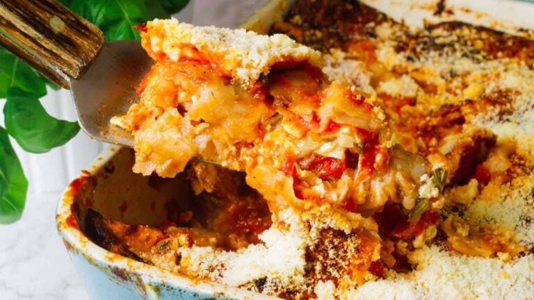 A baked lasagna in a rectangular dish, topped with tomato sauce, basil leaves, and grated cheese. A spatula is lifting a portion from the dish, revealing layers of pasta and filling. The dish is on a light countertop.