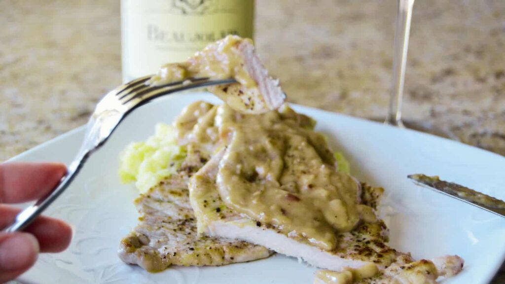 A plate with sliced pork topped with creamy mushroom sauce, a fork holding a piece above, and a wine bottle in the background. Perfect for Moms looking to satisfy picky eaters.