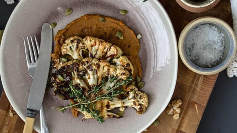 Grilled cauliflower steak served on a bed of purée, garnished with fresh herbs and seeds—a modern twist on Grandma Recipes, perfect for unforgettable side dishes. Presented with a fork and knife on the plate.