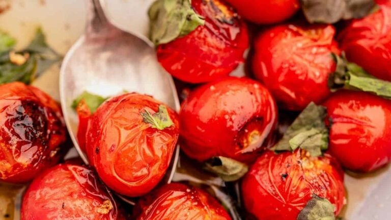 Close-up of roasted cherry tomatoes with charred skins, garnished with wilted basil leaves and a metal spoon in the background.