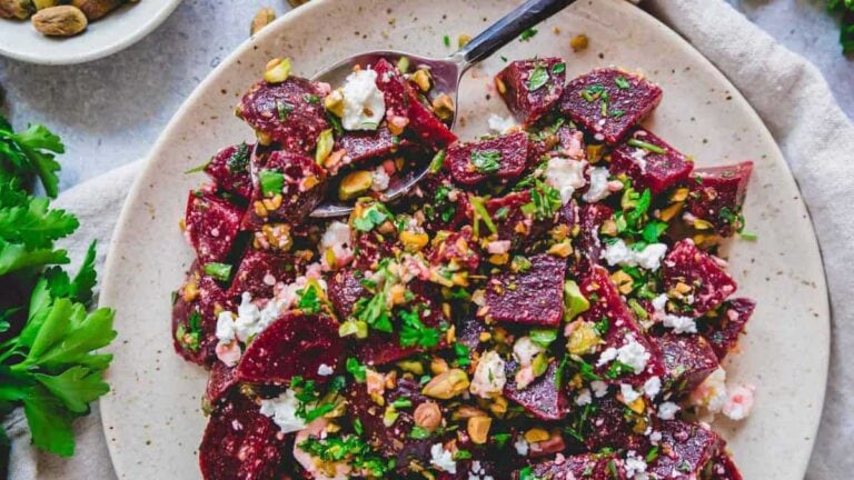 A plate of beet salad topped with crumbled cheese, chopped nuts, and fresh herbs, served with a spoon.