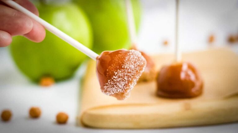 Hand holding a caramel apple pop dusted with sugar; two more caramel pops and green apples rest on a wooden board in the background. A sweet treat perfect for Mother’s Day recipes and fruit lovers alike.