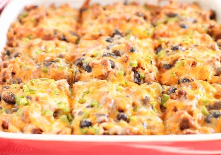 A close-up of a casserole dish containing twelve square portions of baked enchilada casserole topped with melted cheese, black olives, and green onions.