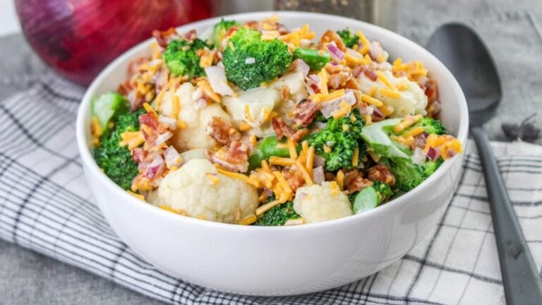 A bowl of broccoli cauliflower salad with shredded cheese and bacon, placed on a checkered cloth.