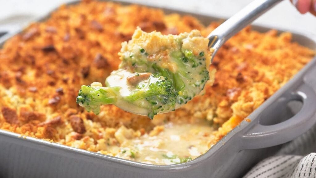 Close-up of a spoon scooping creamy broccoli casserole topped with a golden, crispy layer from a baking dish.