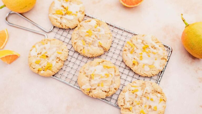 Six glazed citrus cookies are arranged on a cooling rack, with orange slices and whole citrus fruits nearby on a light surface.